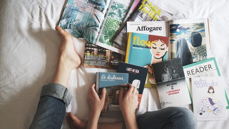 na foto, mãos de uma pessoa segurando um livro e vários livros na cama. quarentena em casa